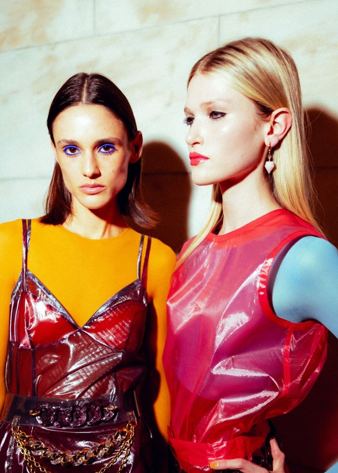 Two models backstage in glossy red and burgundy vinyl looks with bright colour-blocked layers